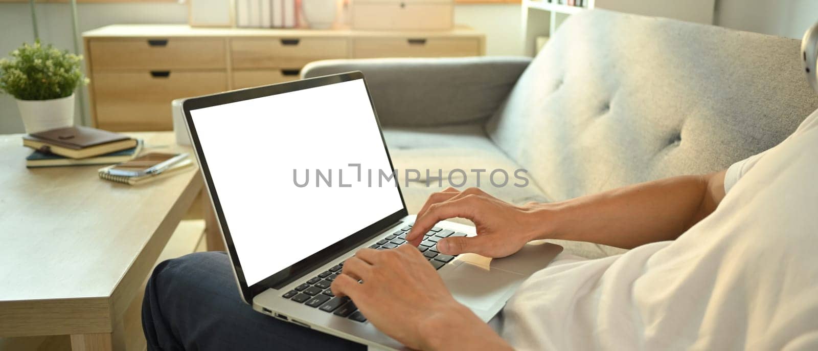 Close up view of man I casual clothes sitting on couch working remotely, surfing internet on laptop by prathanchorruangsak
