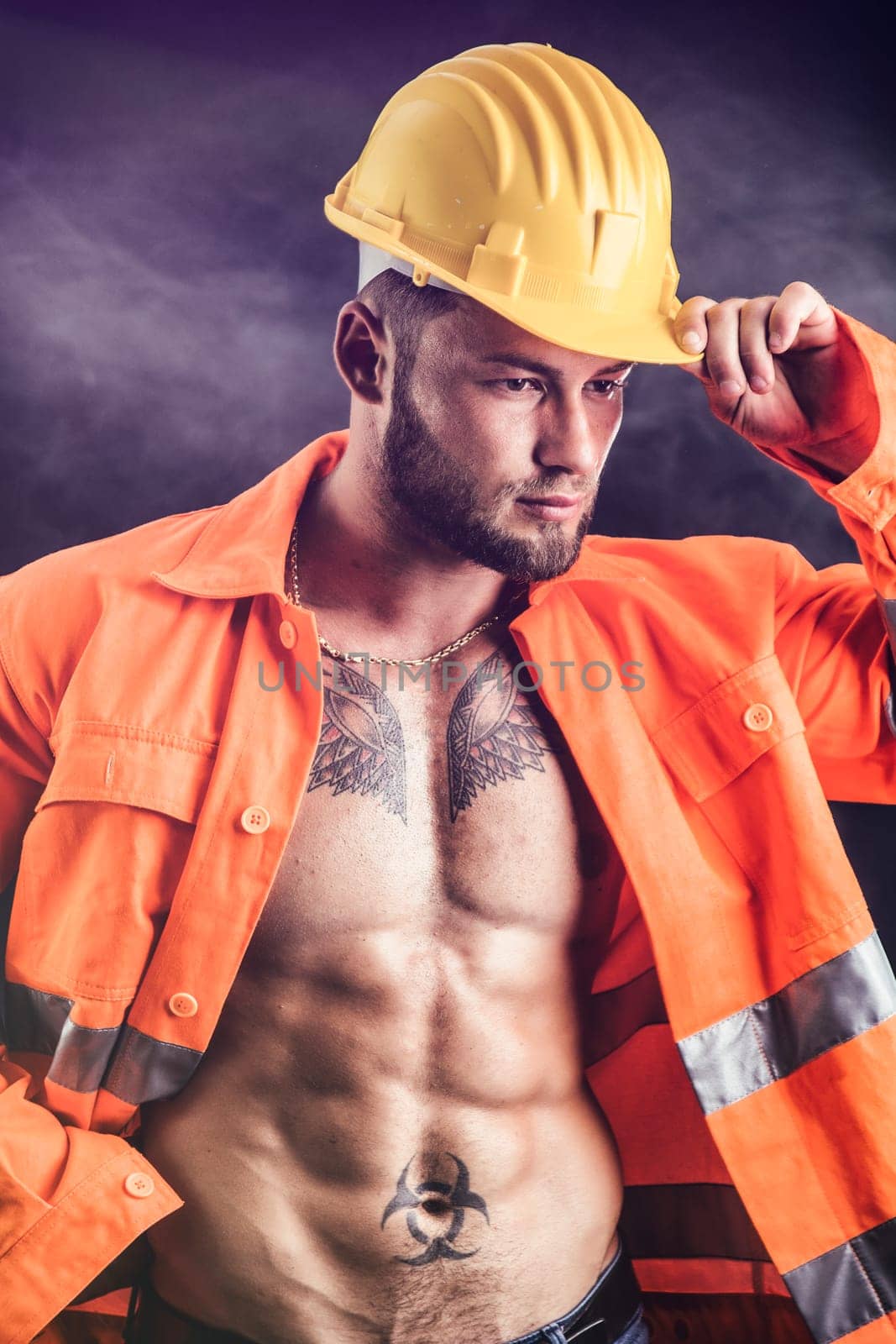 Handsome sexy construction worker with orange suit open on naked torso, wearing yellow hardhat