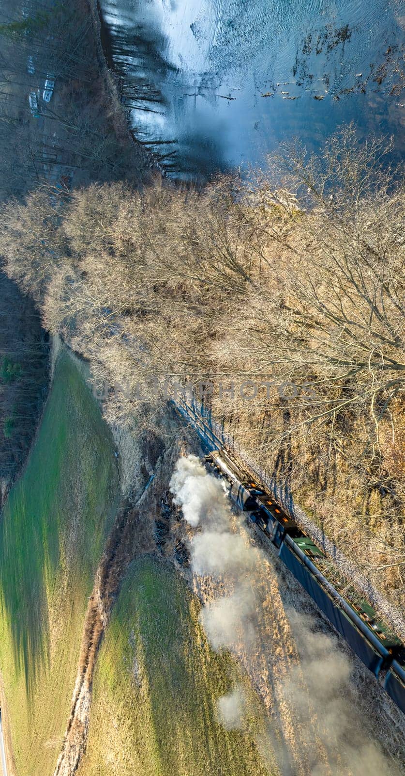 Shirleysburg, Pennsylvania, February 19, 2023 - An Aerial View of a Narrow Gauge Restored Passenger Train Blowing Smoke Traveling Next to a River Thru the Countryside on a Beautiful Sunny Day