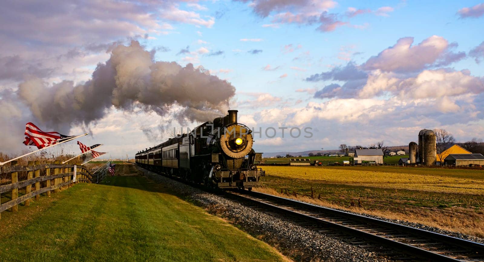 A View of a Classic Steam Passenger Train Approaching, With American Flags Attached to a Fence on a Sunny Autumn Day