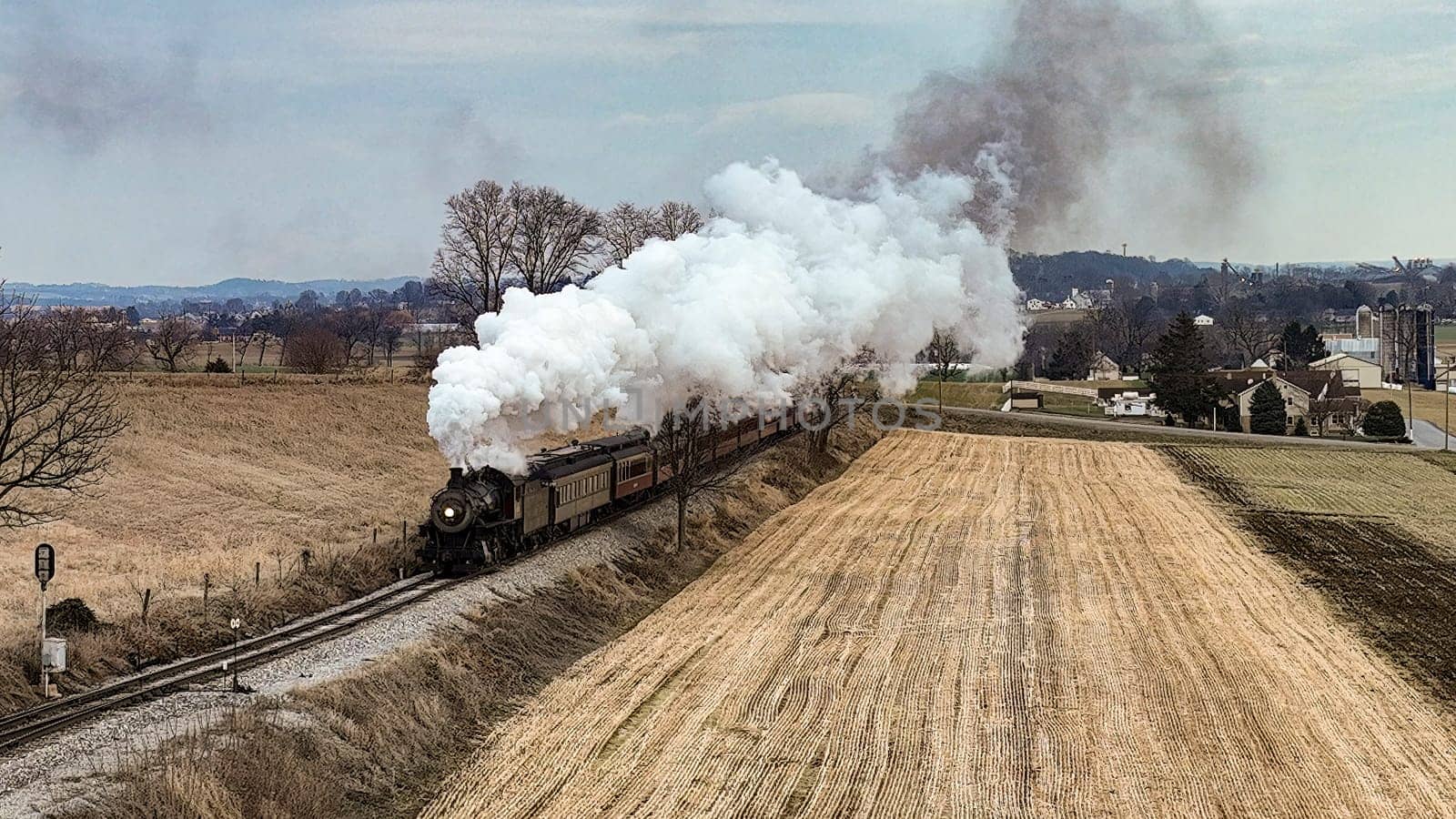 A Drone View of a Steam Locomotive Approaching Traveling Thru Fields and Meadows, Blowing White Smoke on a Winter Day