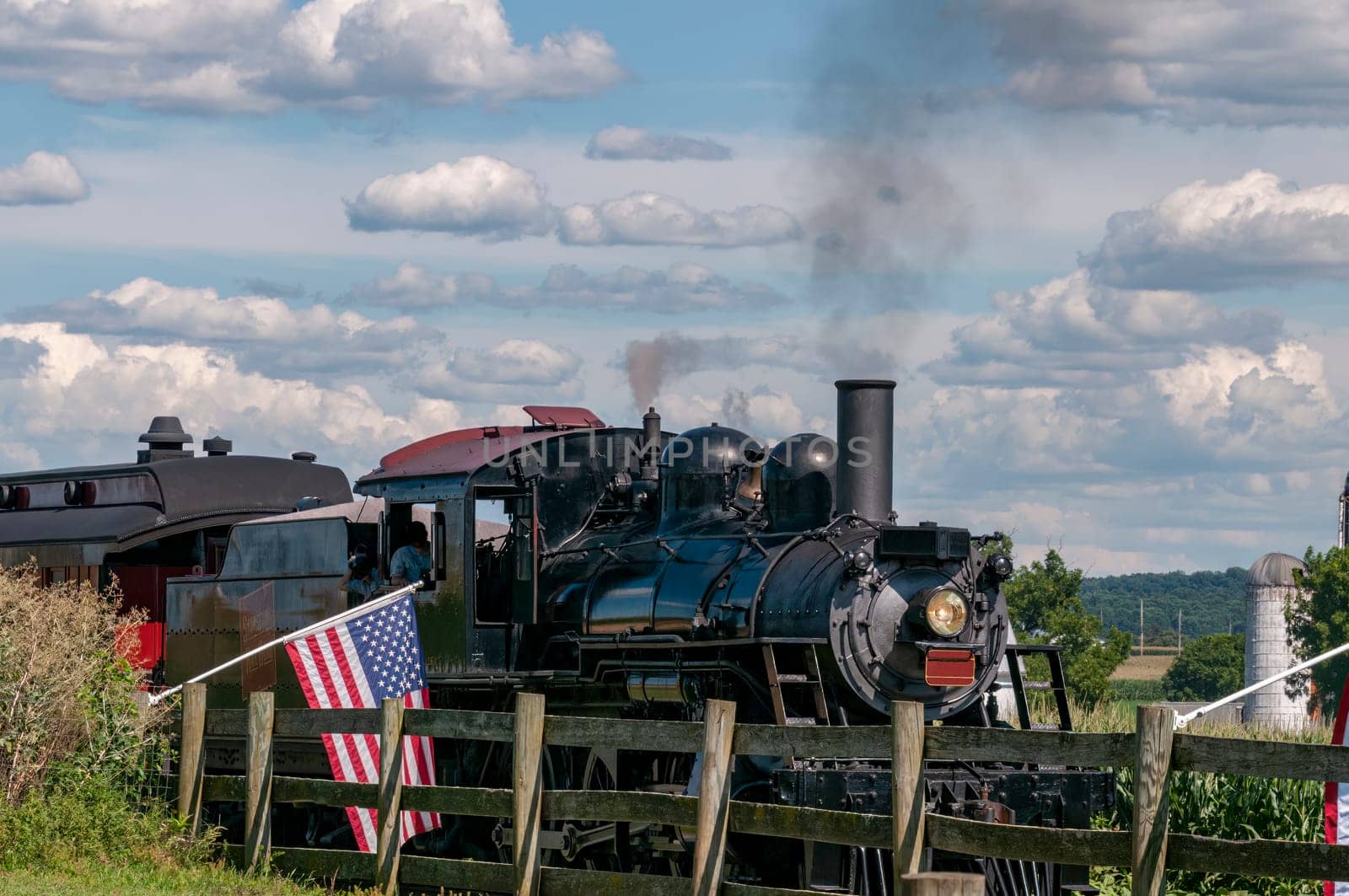 A Restored Antique Steam Passenger Train Approaches a Fence With Gently Waving American Flags on It On a Sunny Summer Day