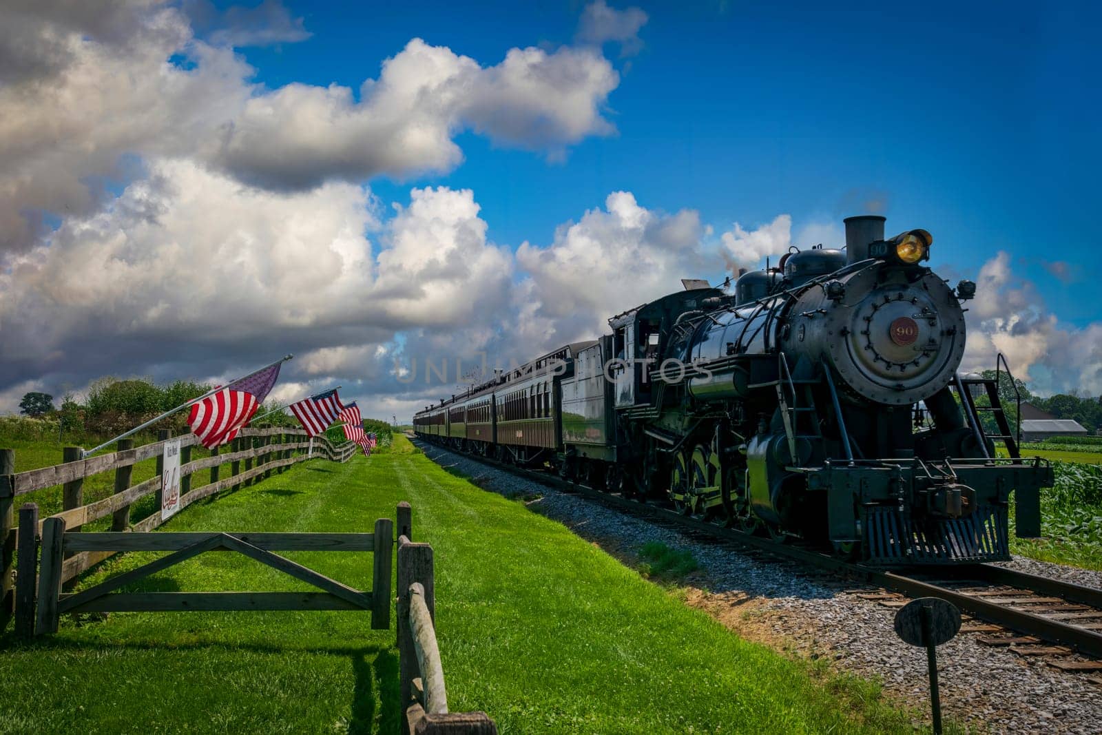 Ronks, Pennsylvania, July 19, 2022 - A Restored Antique Steam Passenger Train Approaches a Fence With Gently Waving American Flags on It On a Sunny Summer Day