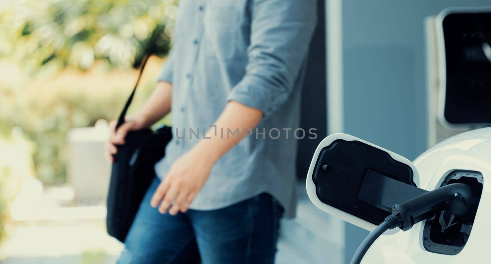 Focus electric car charging at home charging station with blurred progressive man walking in the background. Electric car using renewable clean for eco-friendly concept.