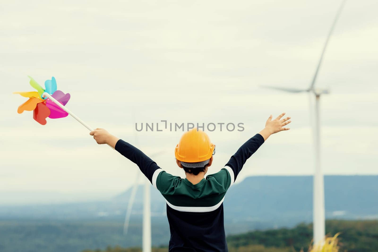 Progressive young asian boy playing with wind pinwheel toy in the wind turbine farm, green field over the hill. Green energy from renewable electric wind generator. Windmill in the countryside concept