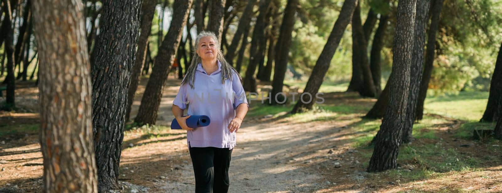 Banner mature elderly woman with grey hair dreadlocks going to workout in park and standing with mat for exercises in green park copy space by Satura86