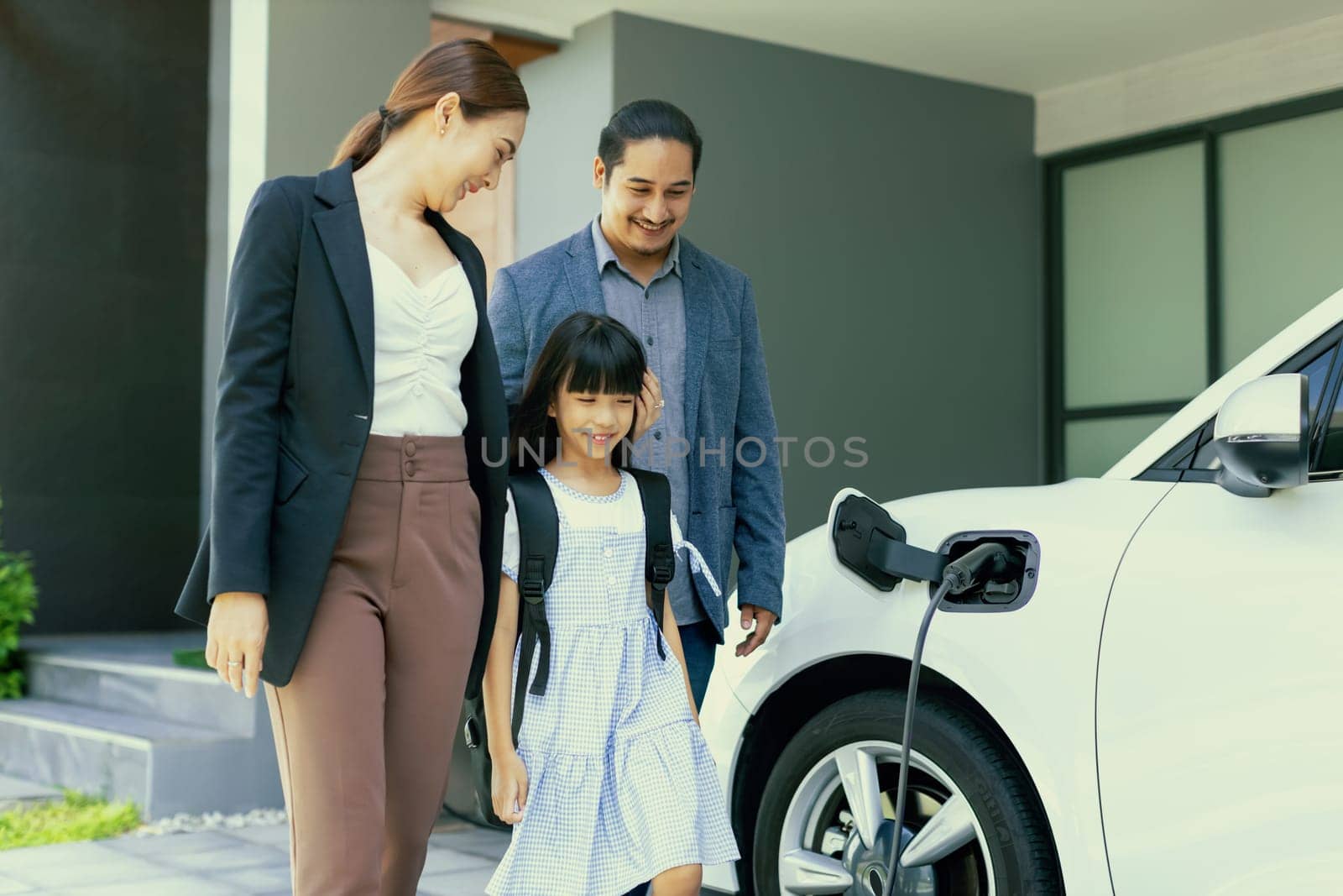 Progressive young parents and daughter with electric vehicle and home charging station. Green and clean energy from electric vehicles for healthy environment. Eco power from renewable source at home.