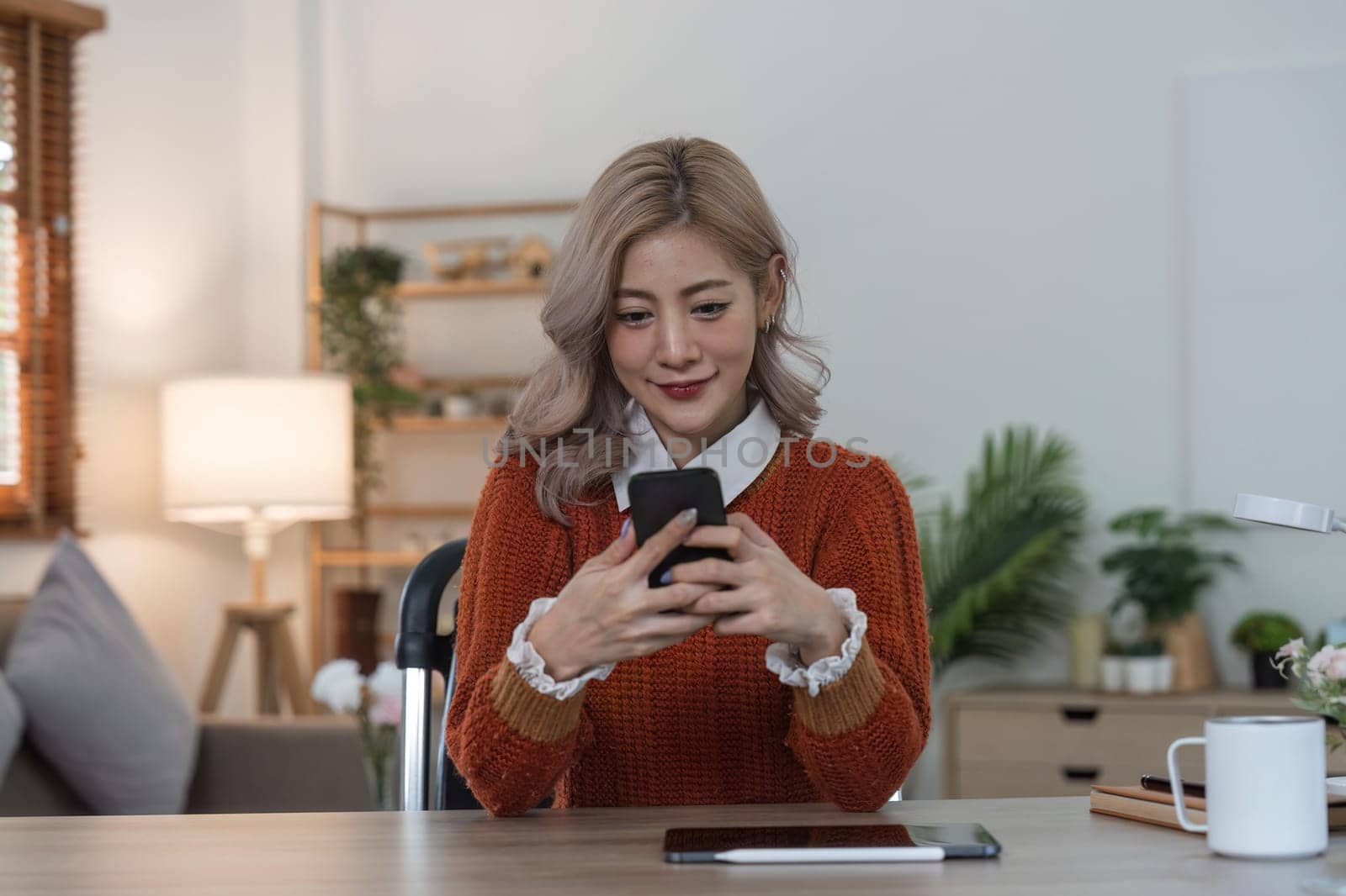 Cute asian woman looking at the smartphone in her hands. Female using phone while sitting down on the desk of her.