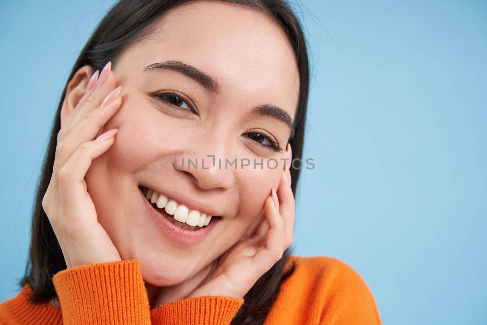 Beauty and skincare. Close up portrait of happy smiling japanese woman, touches her clear, glowing skin, natural healthy face, standing over blue background.