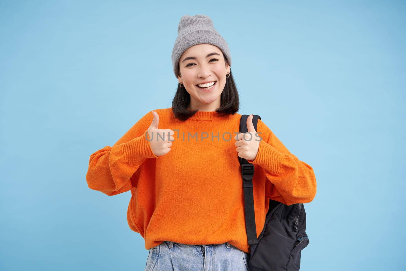 Enthusiastic asian woman in hat, holds backpack, shows thumbs up, approves smth, gives positive feedback, says yes, blue background.