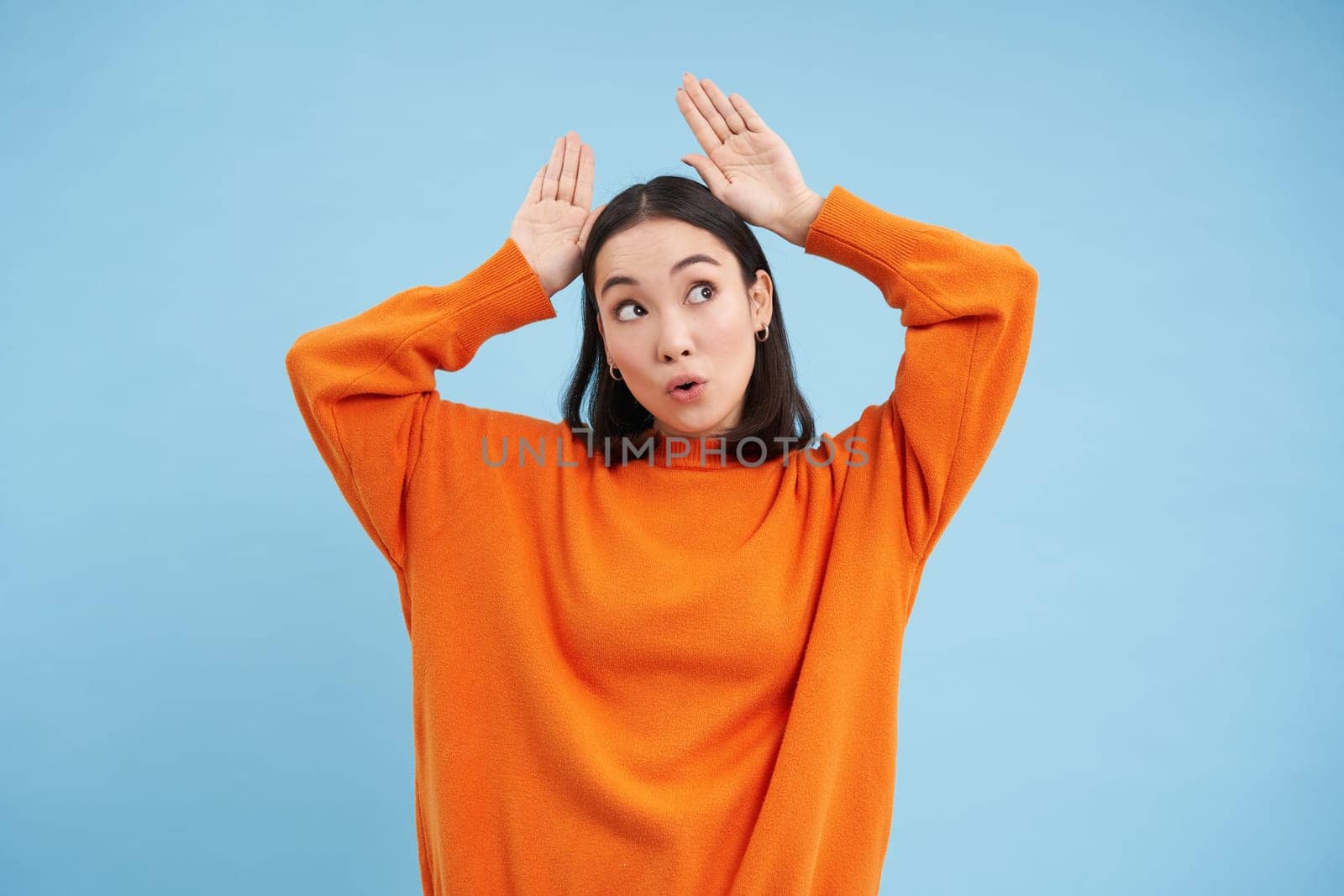 Funny young woman shows ears gesture above head and dancing, looking cute and silly, playing, standing over blue background.