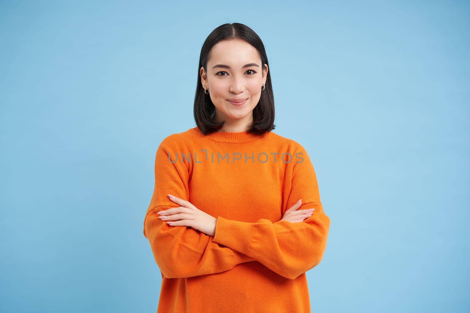 Young asian woman looks with confidence, cross arms on chest and smiles at camera, stands in orange sweatshirt, blue background.