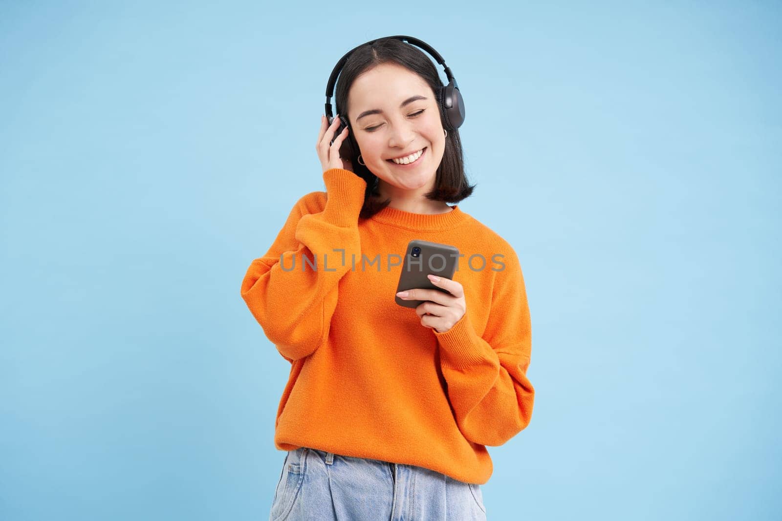 Happy young modern woman with headphones and smartphone, listening to music on mobile phone, enjoys favourite podcast, blue background.