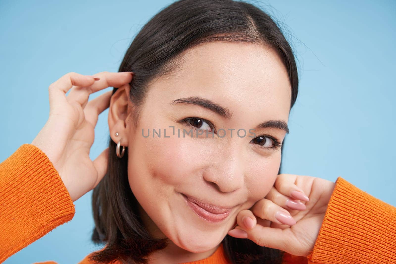 Beauty and skincare. Close up portrait of happy smiling japanese woman, touches her clear, glowing skin, natural healthy face, standing over blue background.