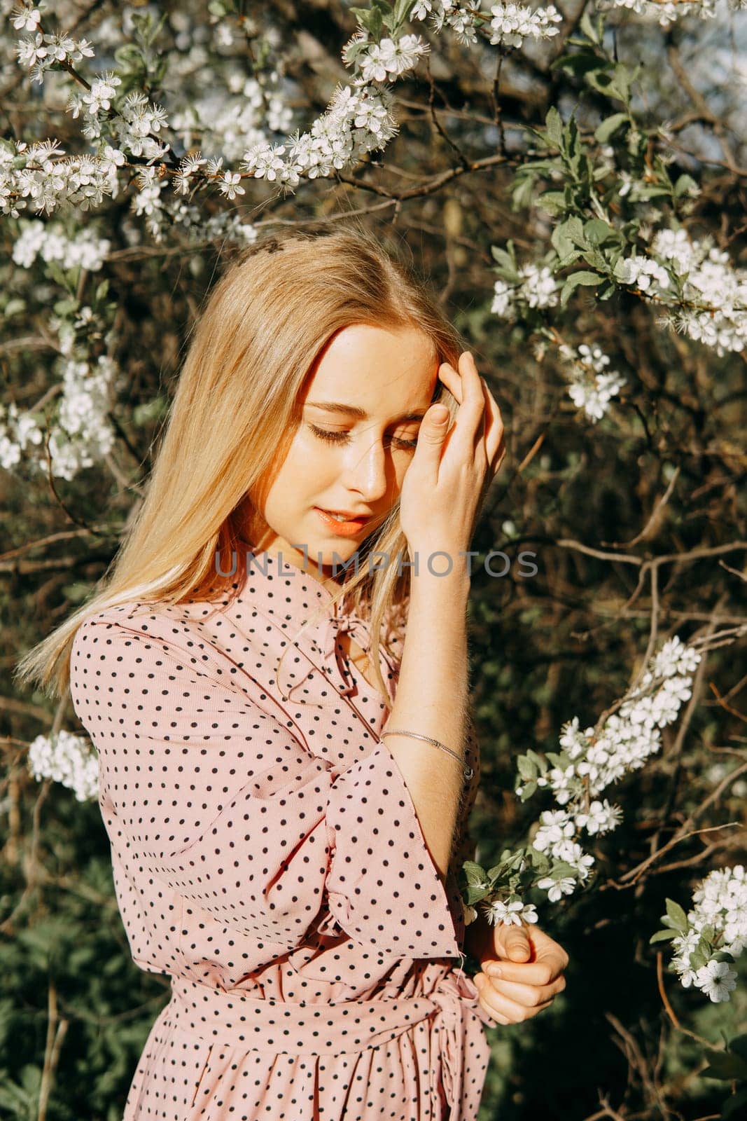 Blonde girl on a spring walk in the garden with cherry blossoms. Female portrait, close-up. A girl in a pink polka dot dress