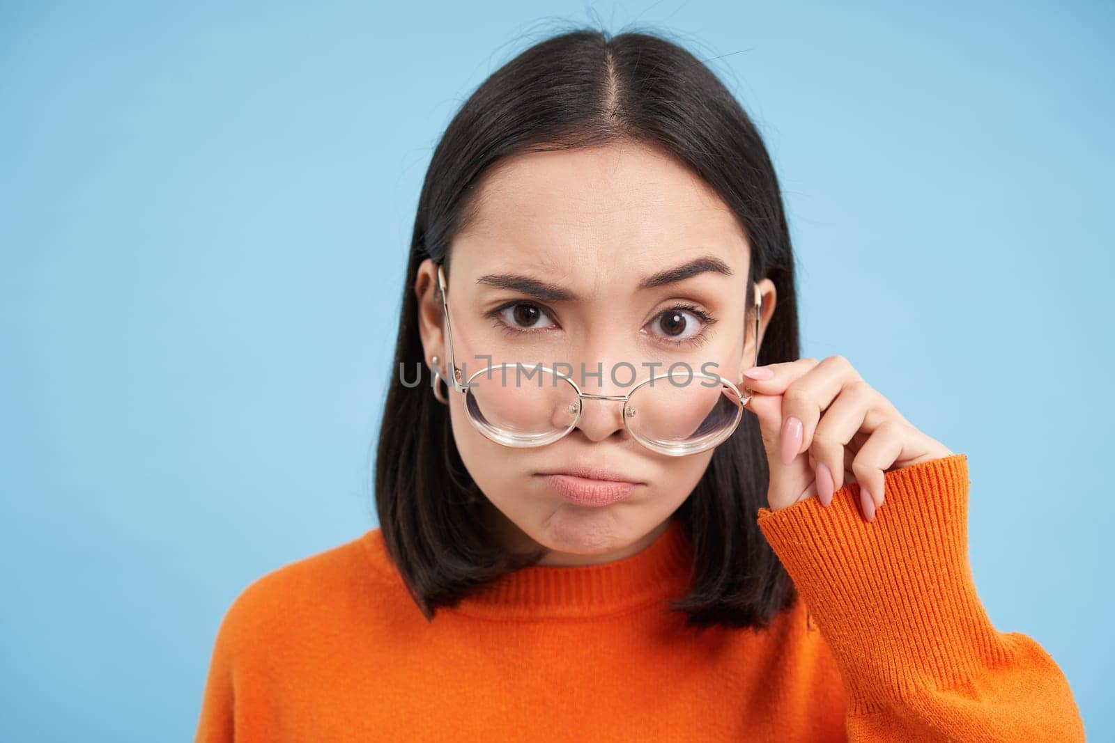 Portrait of asian woman in glasses, stares at camera suspicious, looks with doubt, stands over blue background.