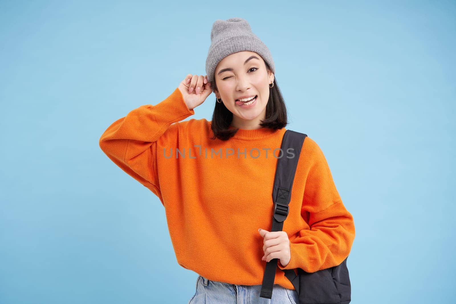 Beautiful modern korean woman in warm hat, holds bag, winks and smiles at camera, blue studio background.