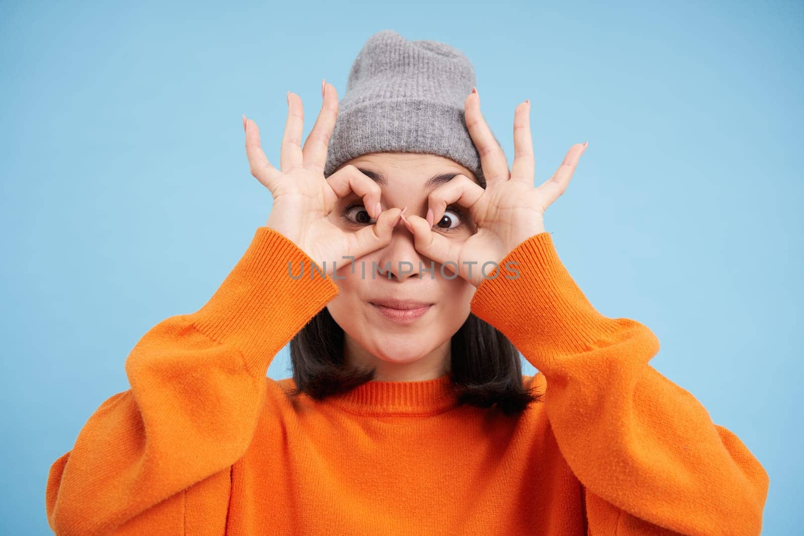 Close up portrait of funny Chinese girl, looks through hand glasses with surprised face expression, standing over blue background.