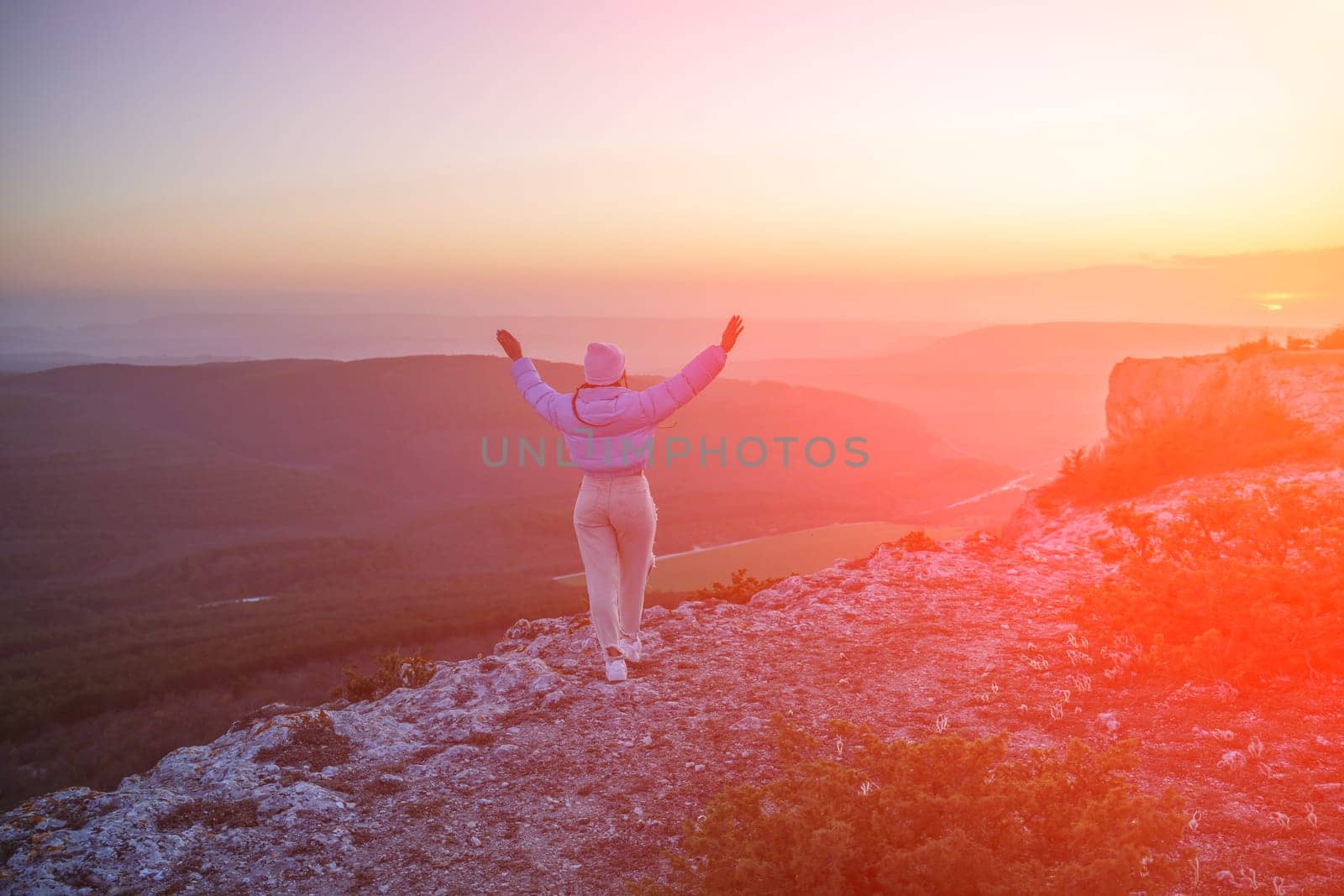Woman hiker open arms on top of sunrise mountain. The girl salutes the sun, wearing a blue jacket, white hat and white jeans. Conceptual design. by Matiunina