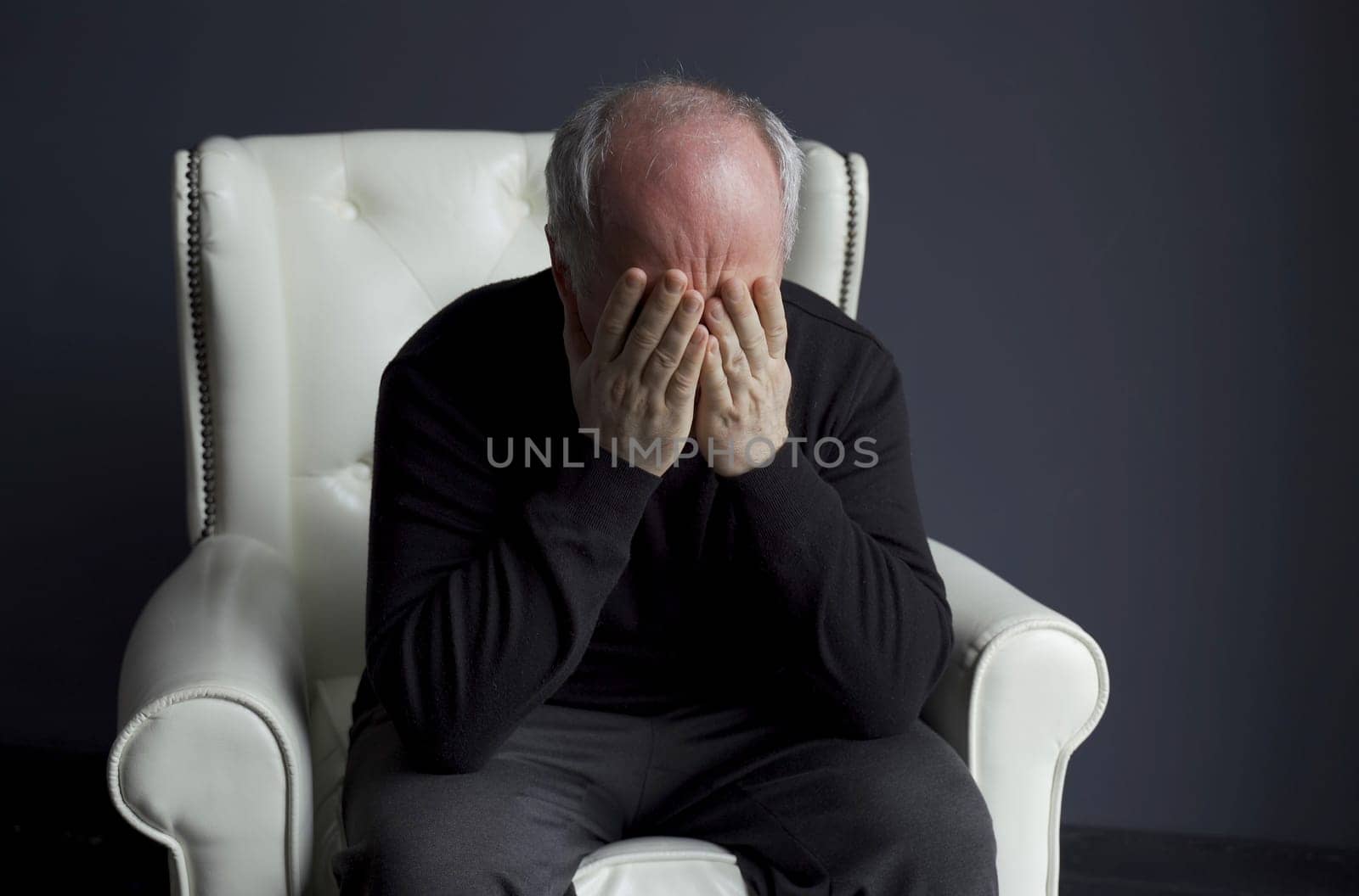 A man in a black turtleneck is sitting in a white chair, rubbing his face with his hands, holding his head. Depression, headache. Medical concept.