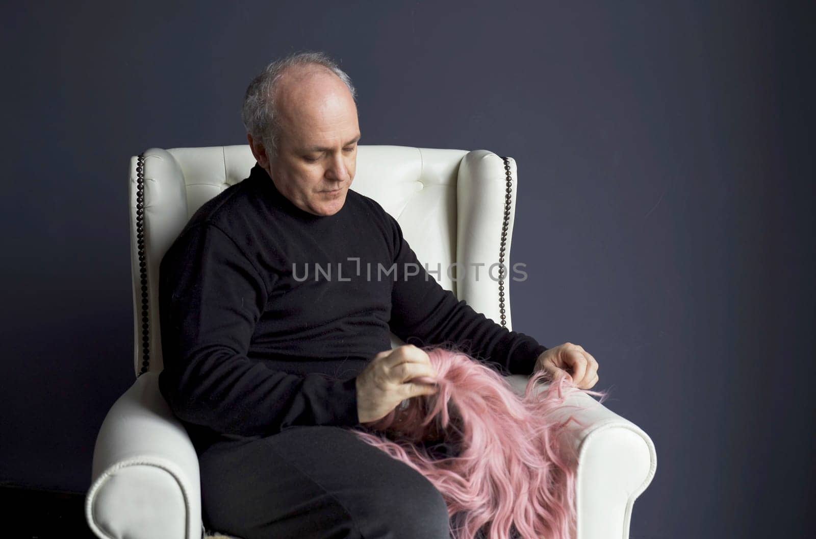 A middle-aged man sits in a chair and looks at a pink wig. Baldness problem.