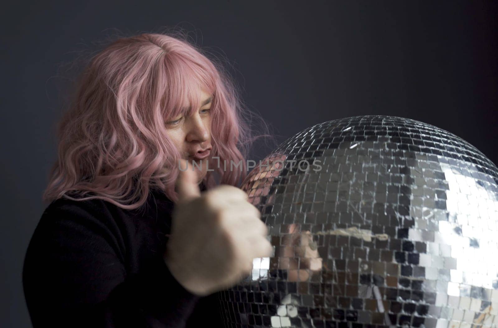 A man in a black turtleneck and a pink wig is dancing, holding a mirrored disco ball in his hand, showing a hand gesture class. Close-up.