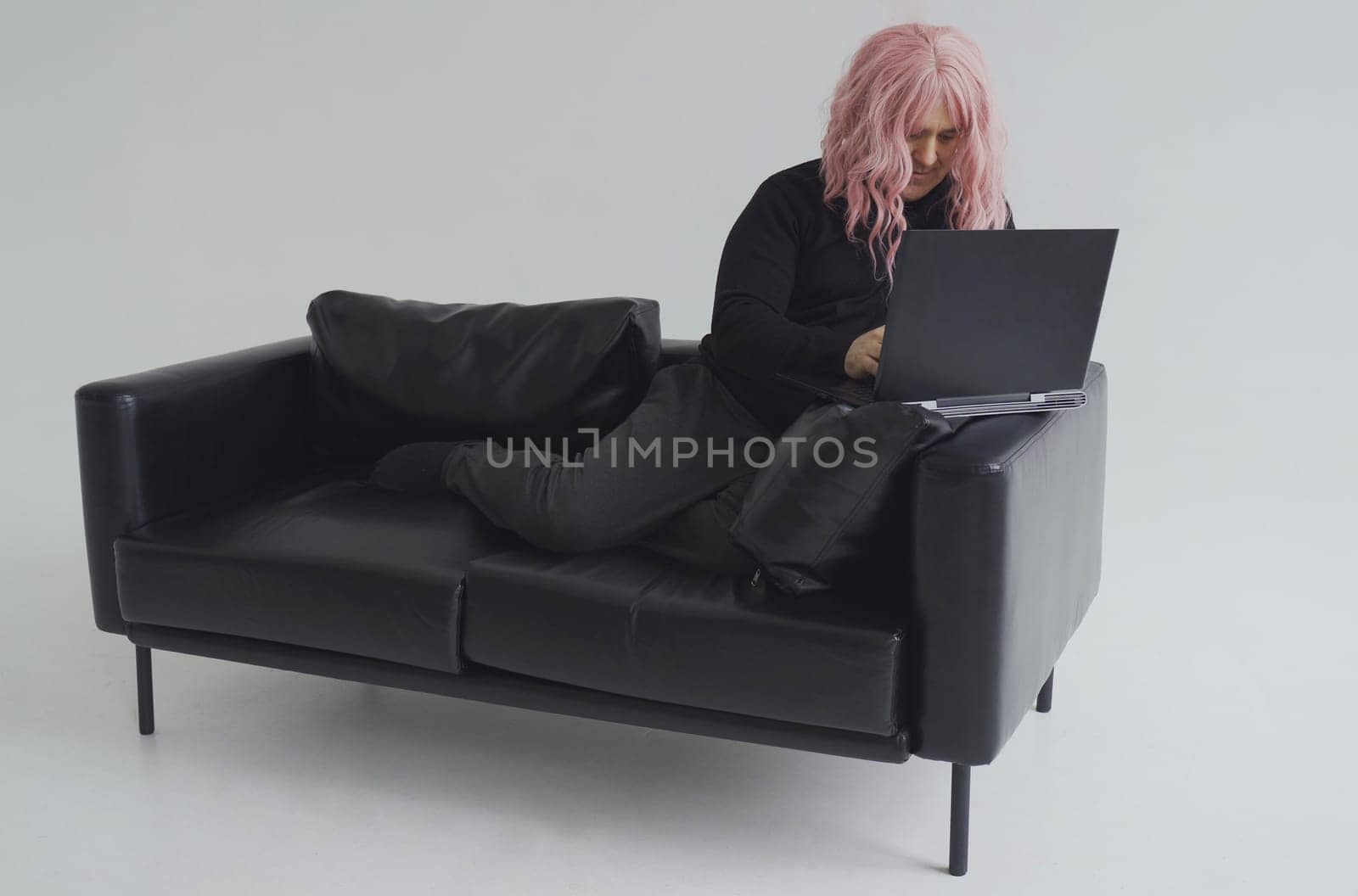 Portrait of a man in a pink wig, sitting on a sofa, typing on a laptop. White background.