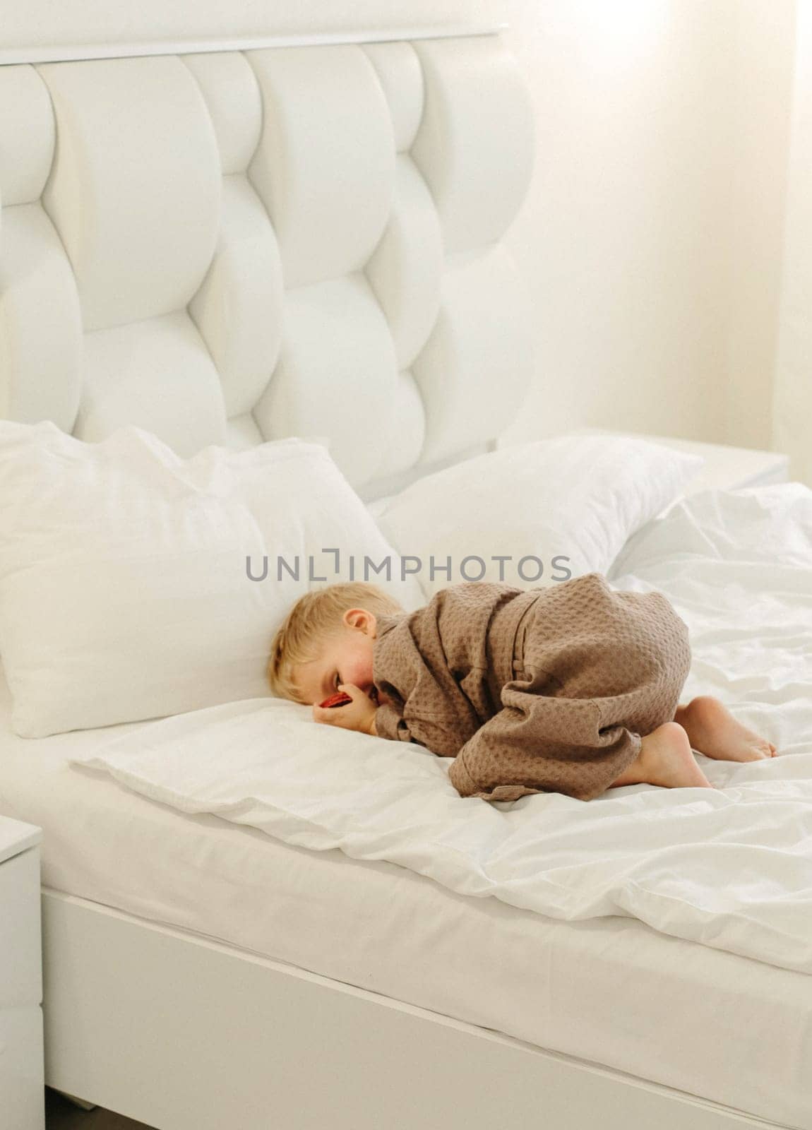 A boy in a brown bathrobe lies on a white bed, peeping through his hands. Vertical frame.