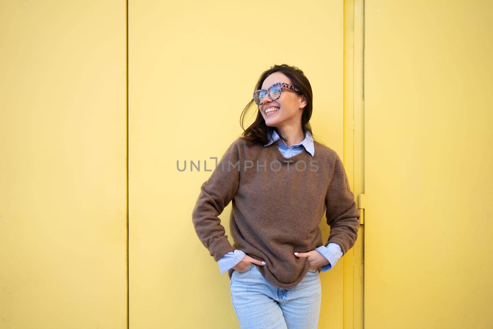 Happy woman in glasses outdoor isolated on yellow color background. Positive people concept. Smiling girl looking side, hands in pocket, dressed sweater and jeans