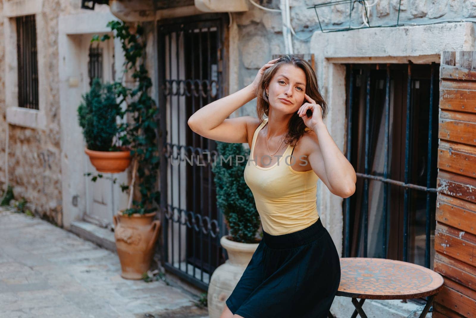 Girl Tourist Walking Through Ancient Narrow Street On A Beautiful Summer Day In MEDITERRANEAN MEDIEVAL CITY, OLD TOWN BUDVA, MONTENEGRO. Young Beautiful Cheerful Woman Walking On Old Street At Tropical Town. Pretty Girl Looking At You And Smiling by Andrii_Ko