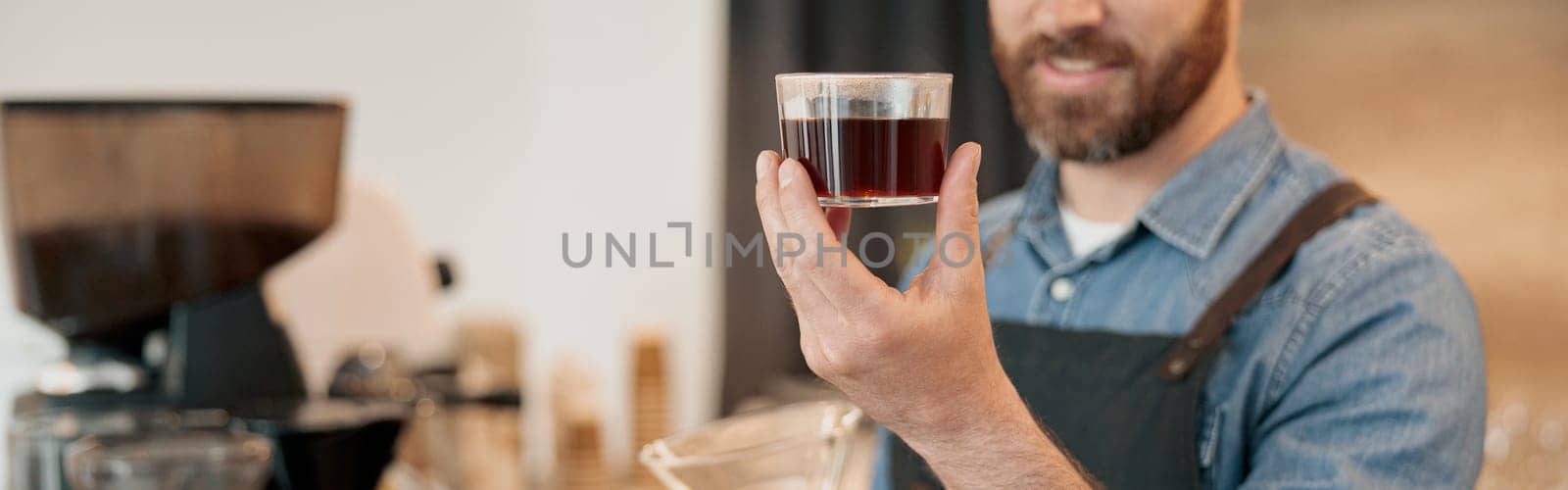 Close up of smiling barista wearing apron appraisingly looks at a glass of filter coffee