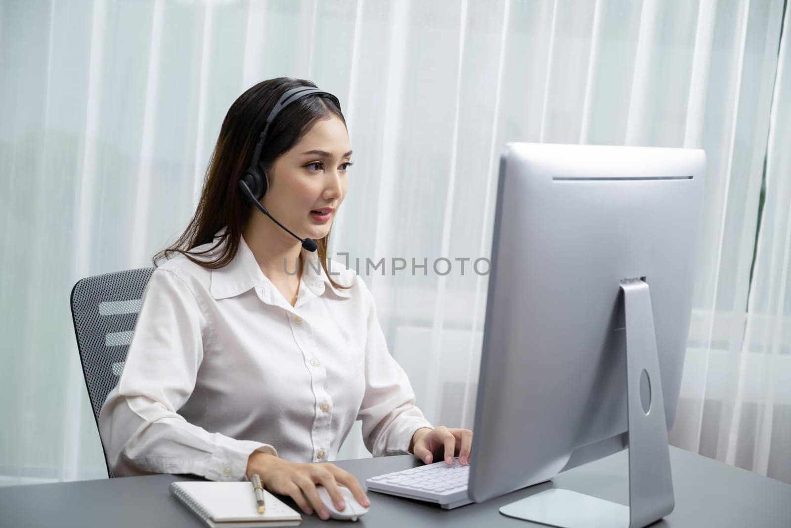 Enthusiastic asian call center with headset and microphone working on her laptop by biancoblue