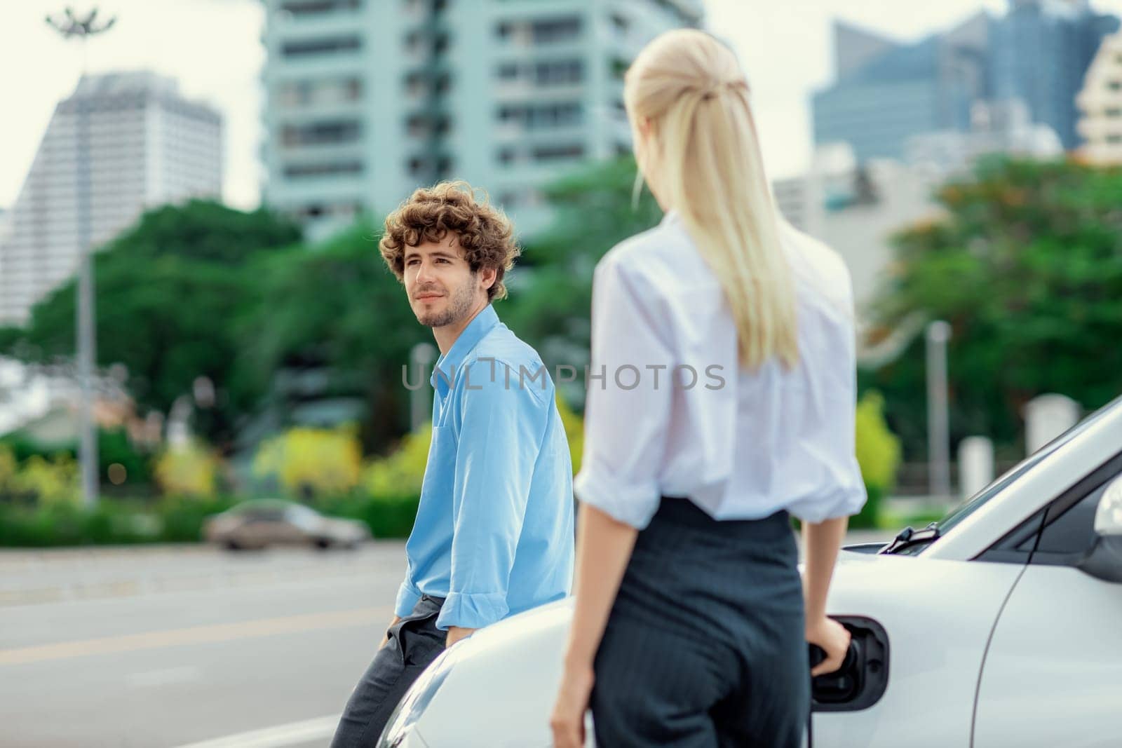 Progressive businessman and businesswoman with electric car parking and connected to public charging station before driving around city center. Eco friendly rechargeable car powered by clean energy.