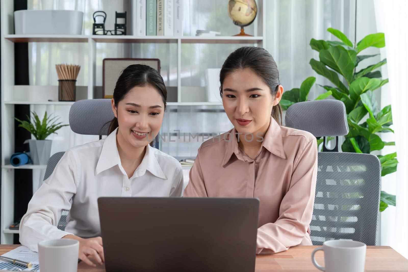 Two young office lady colleagues collaborating in modern office workspace, engaging in discussion and working together on laptop, showcasing their professionalism as modern office worker. Enthusiastic