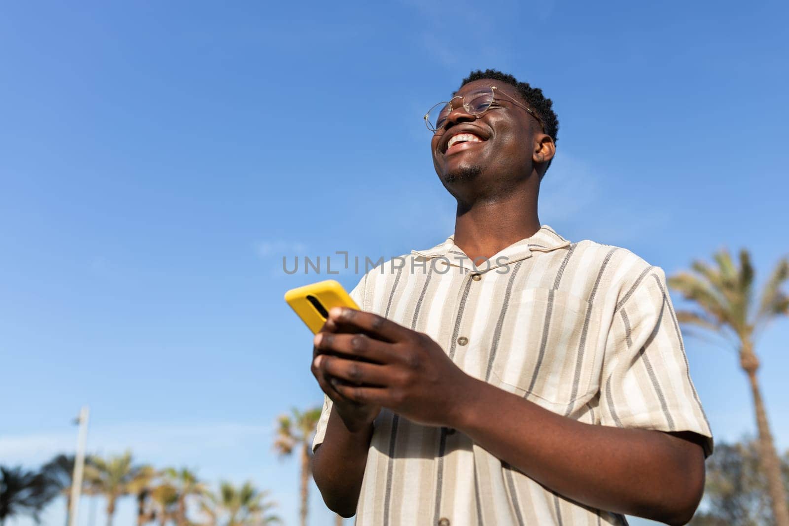 Happy black man laughing outdoors using mobile phone. Blue sky background. Copy space. by Hoverstock