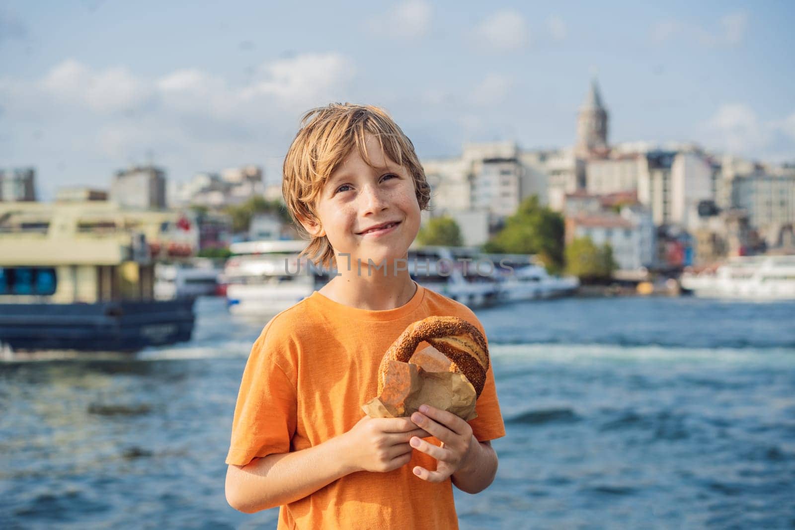 Boy in Istanbul having breakfast with Simit and a glass of Turkish tea. Glass of Turkish tea and bagel Simit against golden horn bay and the Galata Tower in Istanbul, Turkey. Turkiye. Traveling with kids concept.