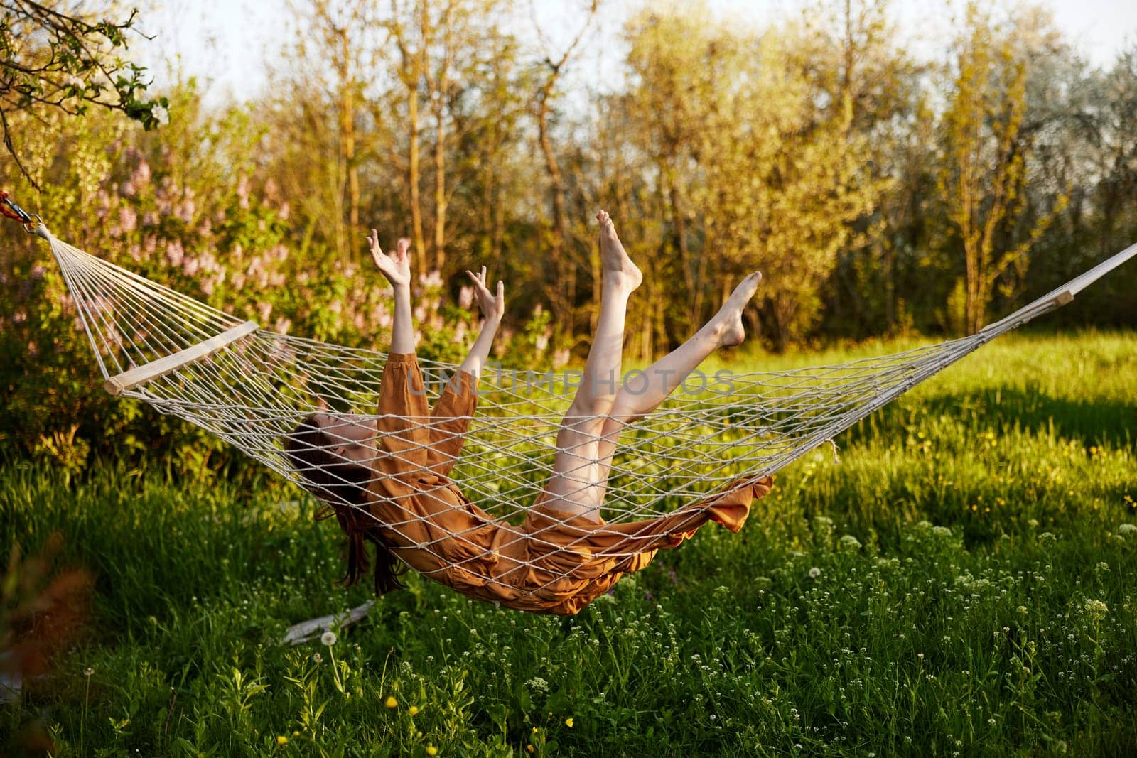 a funny woman is resting in nature lying in a mesh hammock in a long orange dress lifting up her arms and legs. High quality photo