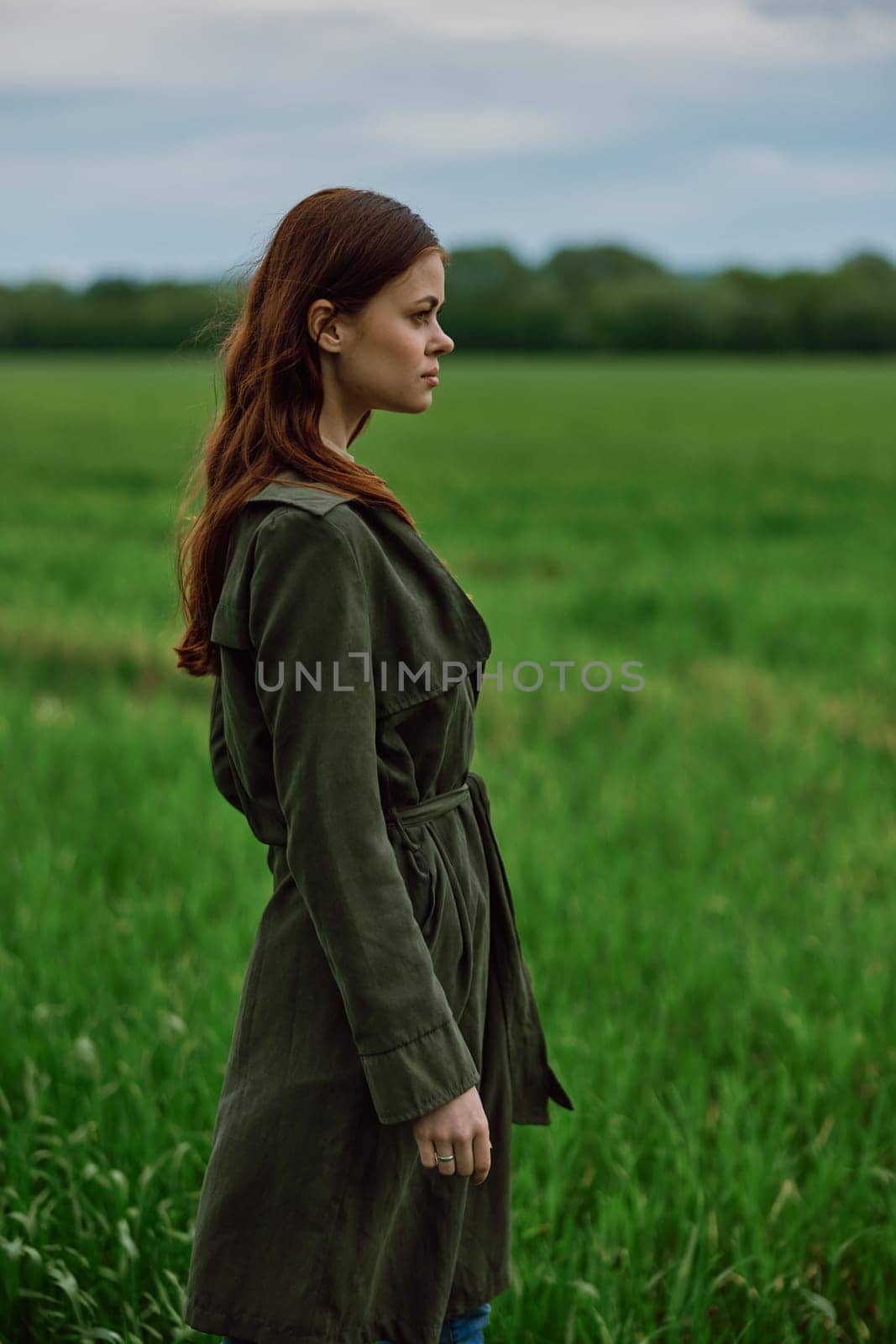 a beautiful, red-haired girl in a raincoat stands in a field in the spring in rainy weather by Vichizh