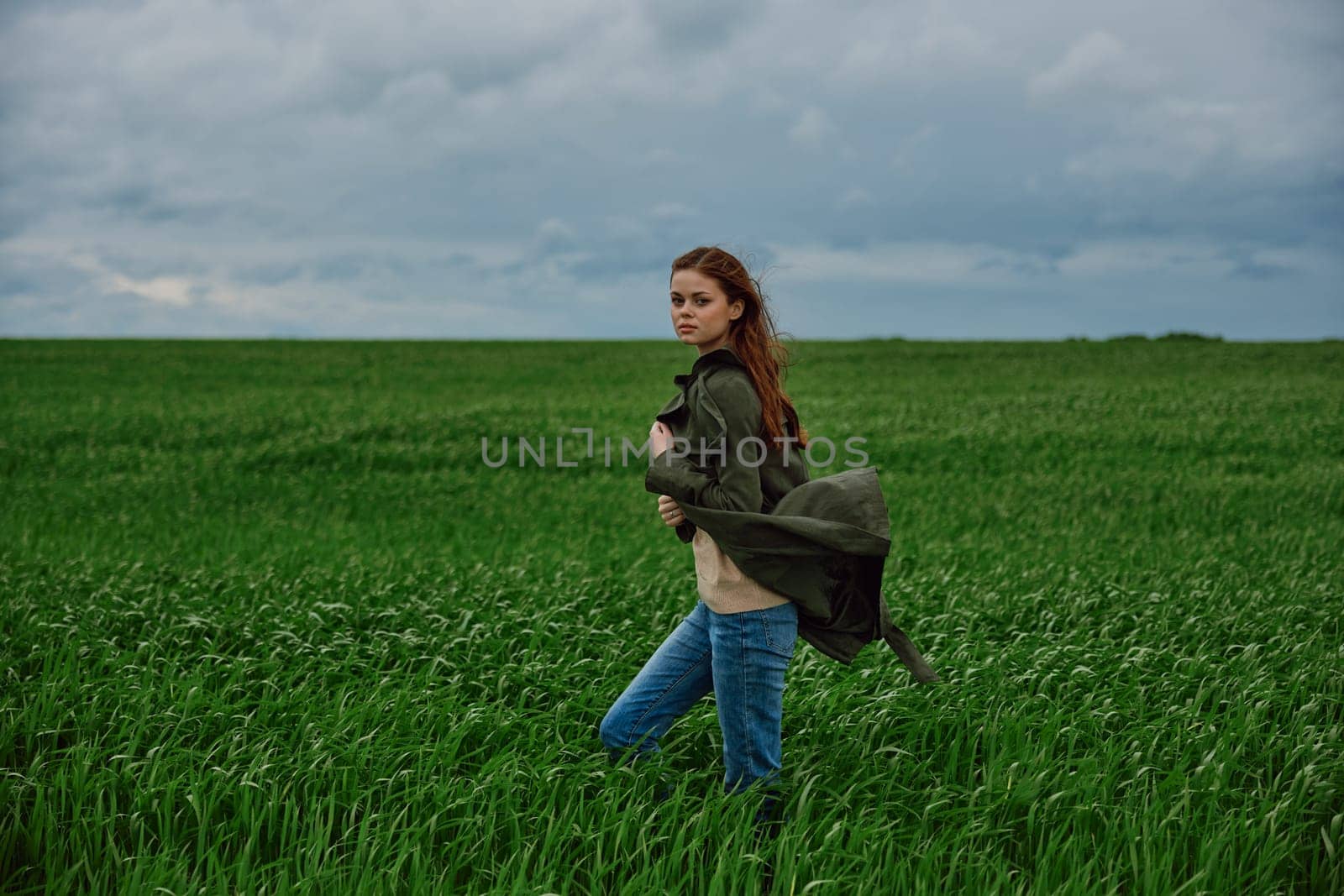 a red-haired woman stands in a green field in rainy, cold weather, holding a raincoat in the wind. High quality photo