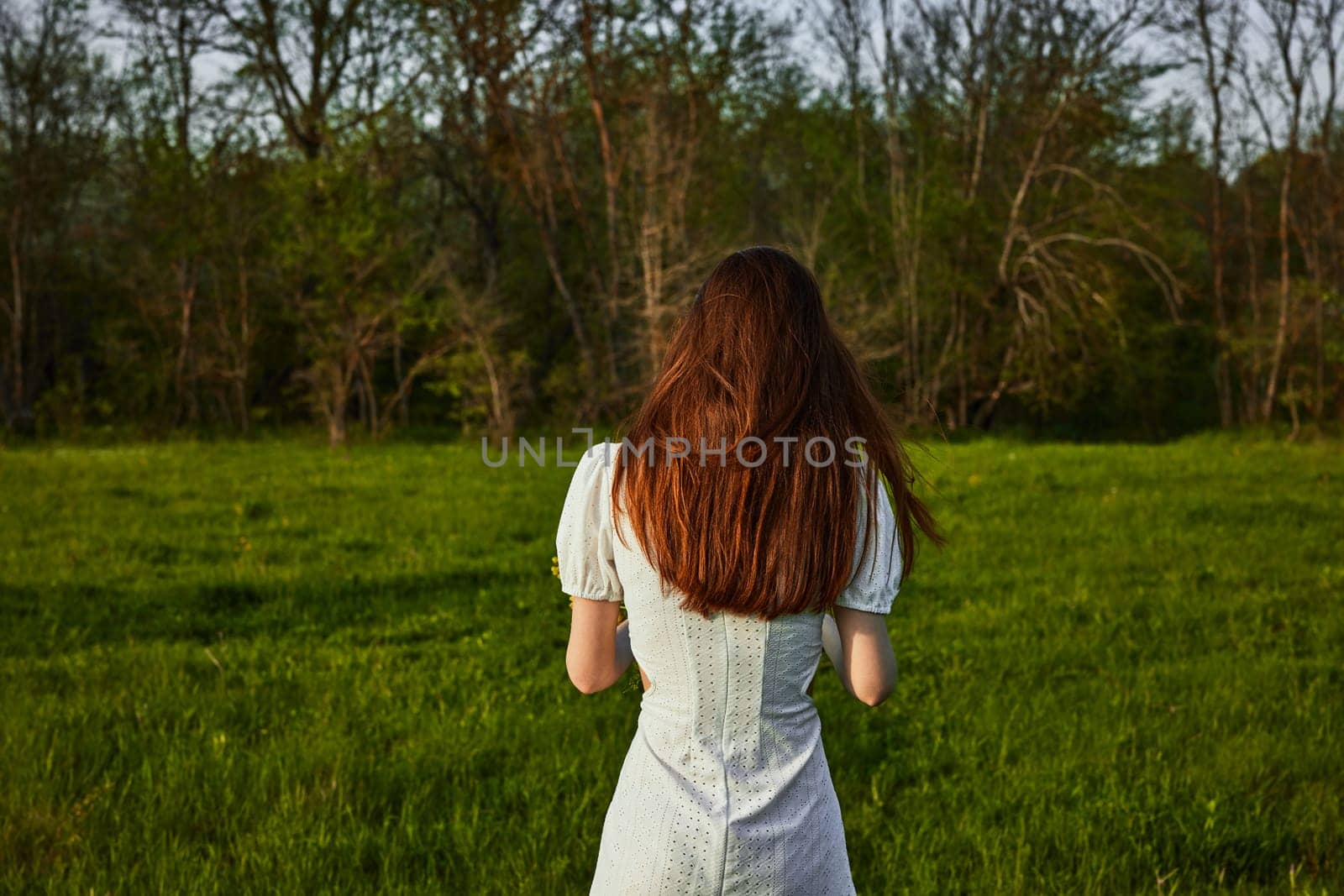 a woman with long red hair in a white dress stands in a green field at sunset with her back to the camera by Vichizh
