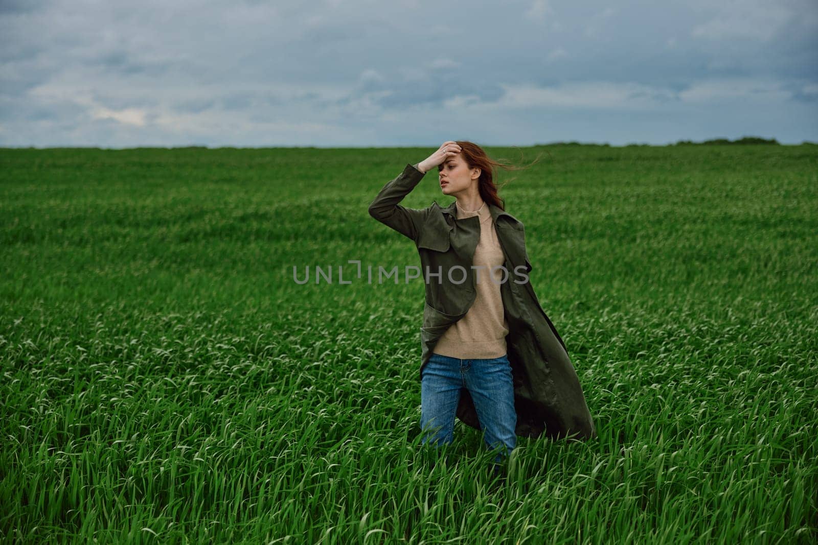 a woman in a dark coat stands in a green field against a cloudy sky holding her hair in the wind by Vichizh