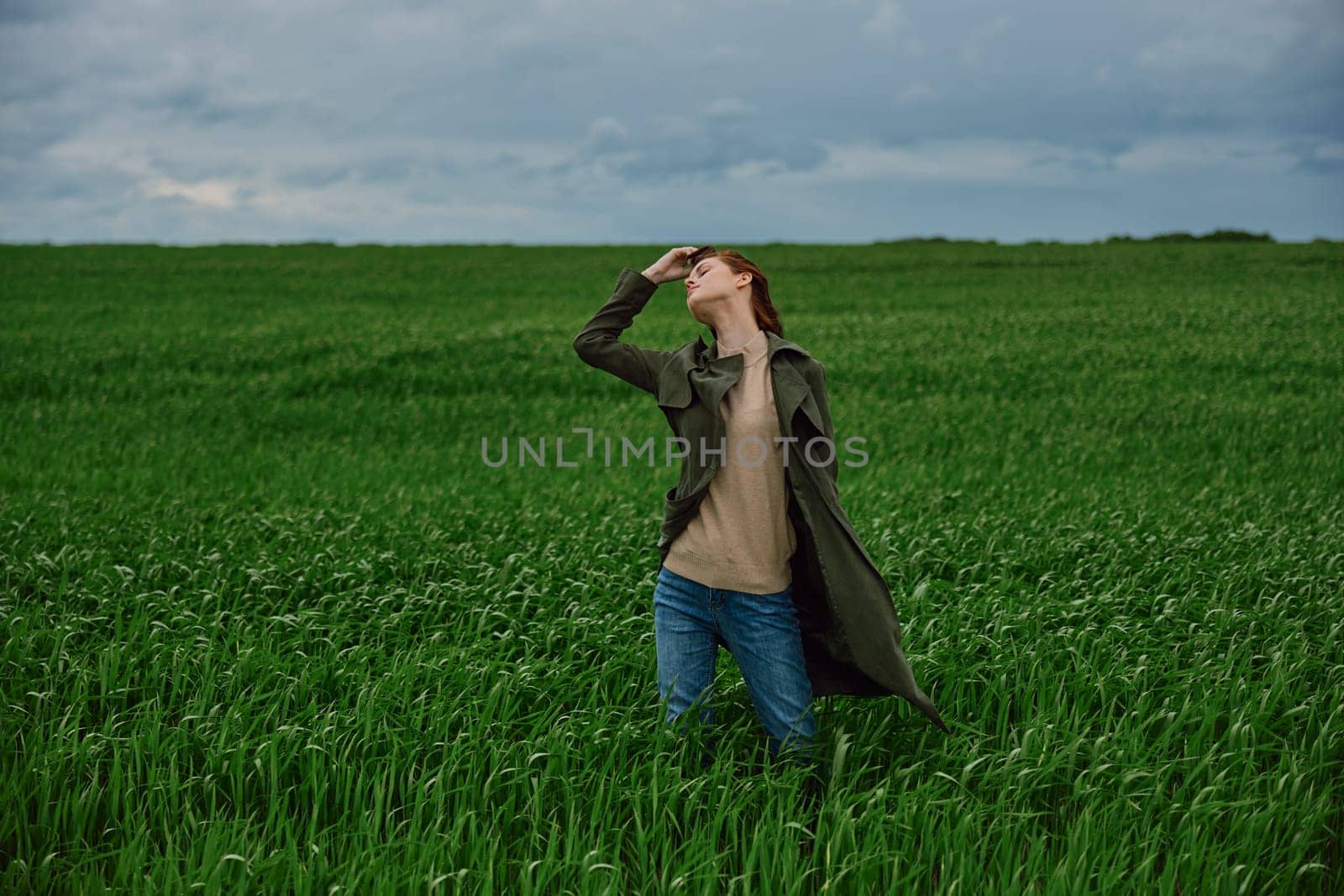 a woman in a dark coat stands in a green field against a cloudy sky holding her hair in the wind. High quality photo