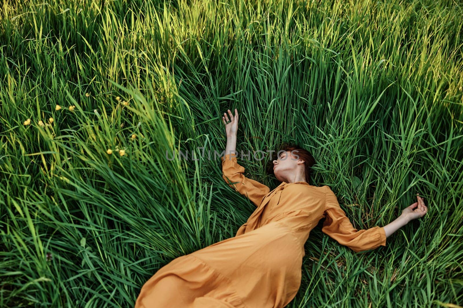 a sweet, calm woman in an orange dress lies in a green field with her arms outstretched and her eyes closed, enjoying the silence and peace. Horizontal photo taken from above. High quality photo