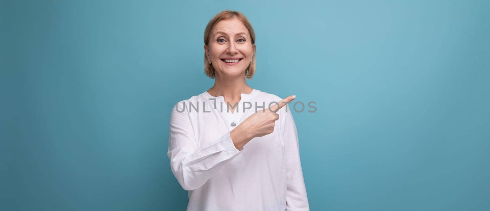 stylish blond middle-aged woman in a white blouse demonstrates something on a studio background with copyspace.