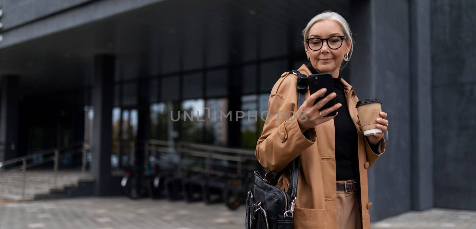 a mature adult female director with gray hair during a break with a phone stands against the backdrop of a business center.