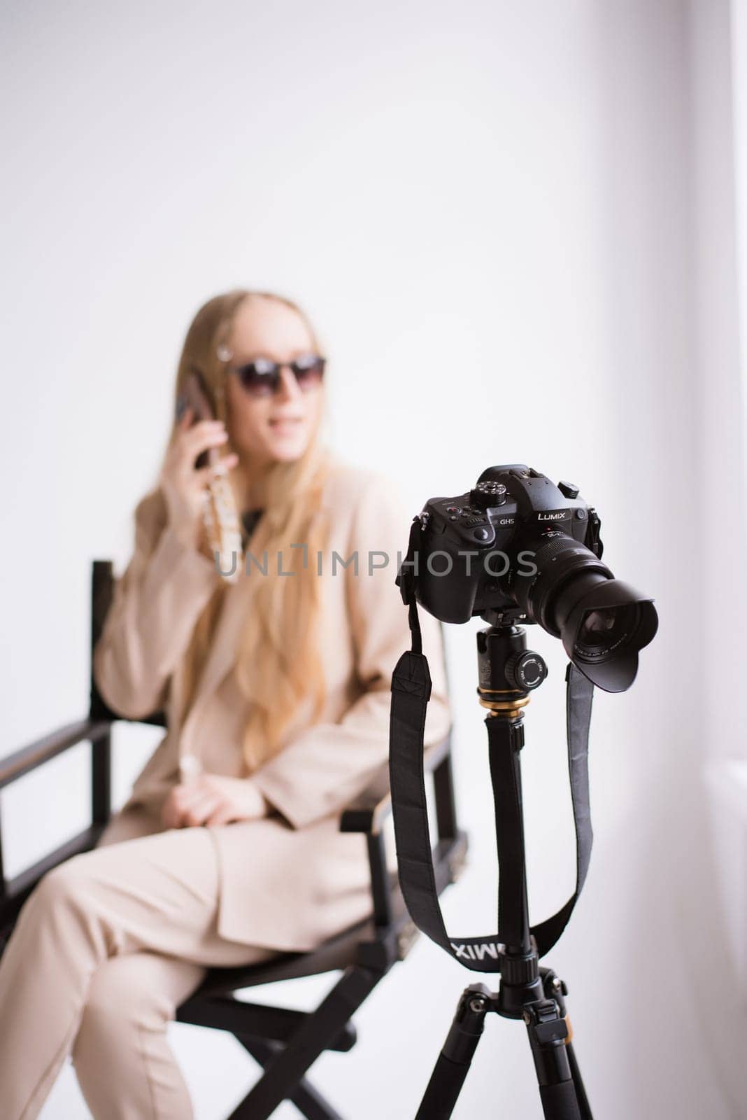 A woman videographer, a blonde with a camera and phone, happy talking on the phone and smiling in the studio. Wearing a formal nude pantsuit on a white isolated background