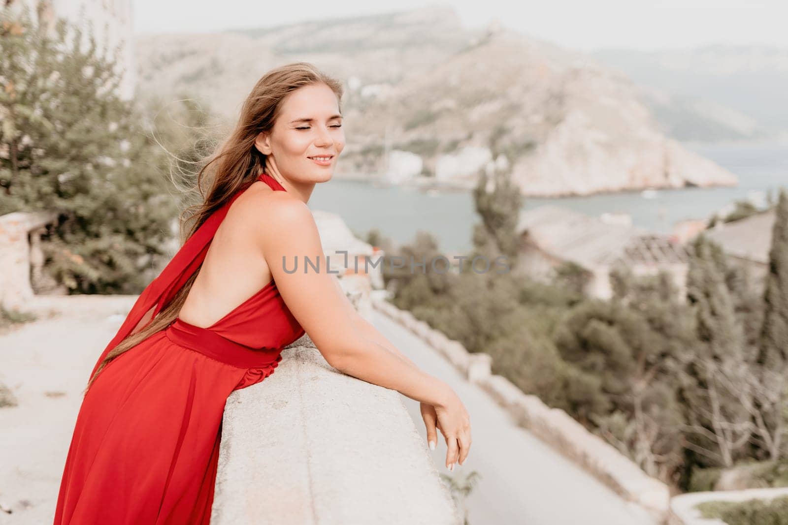 Side view a Young beautiful sensual woman in a red long dress posing on a volcanic rock high above the sea during sunset. Girl on the nature on overcast sky background. Fashion photo