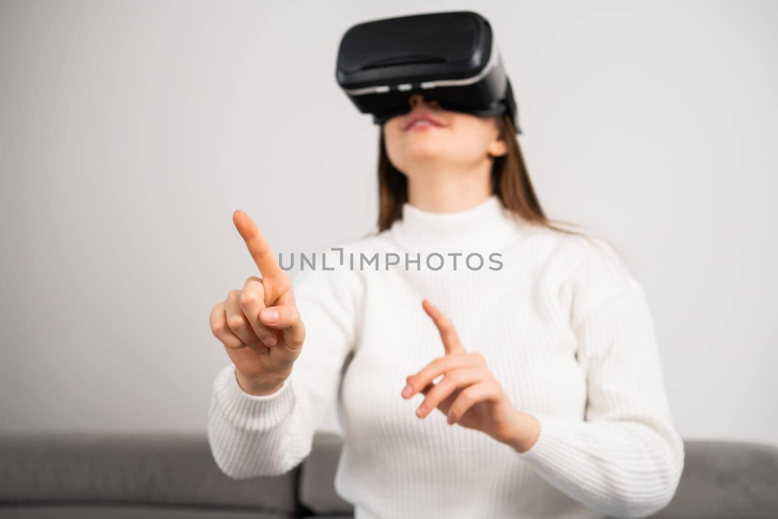 Curious housewife in VR goggles swipes virtual interface to choose game. Young woman rests on couch tasting modern innovations at home