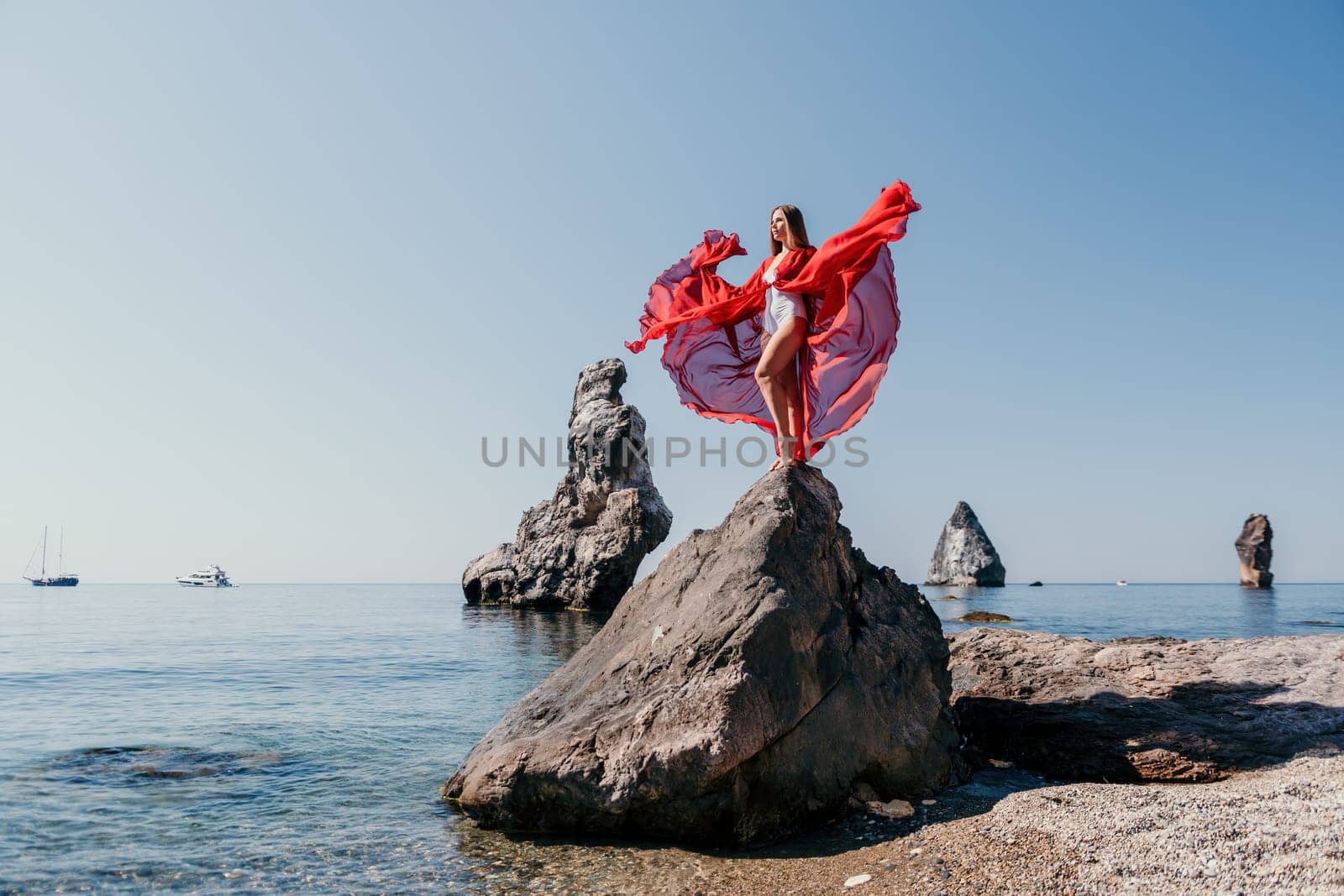 Woman travel sea. Young Happy woman in a long red dress posing on a beach near the sea on background of volcanic rocks, like in Iceland, sharing travel adventure journey by panophotograph