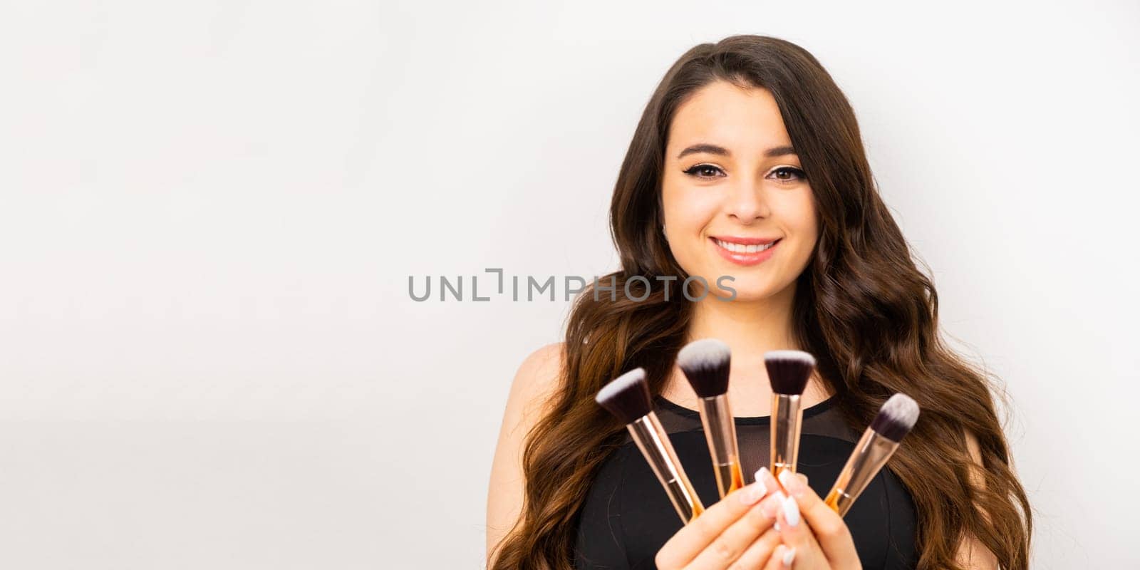 A smiling brunette woman holding makeup brushes on the white background.